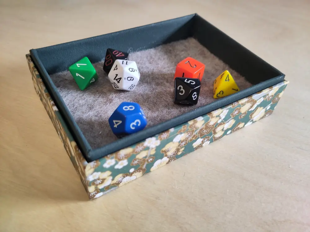 Image of dice box with some dice in it; the box is places into the lid and sticks for a few millimeter out above the edge of the lid.